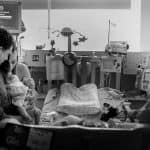a group of women standing around a baby in a hospital bed.