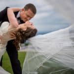 a bride and groom kissing with a veil blowing in the wind.