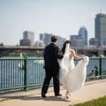 a bride and groom walking on a sidewalk by the water.