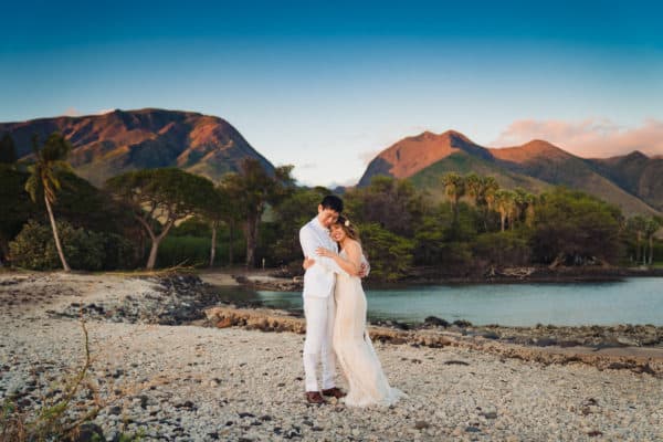 a bride and groom standing on a beach with mountains in the background.