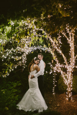 a bride and groom standing under a tree covered in fairy lights.
