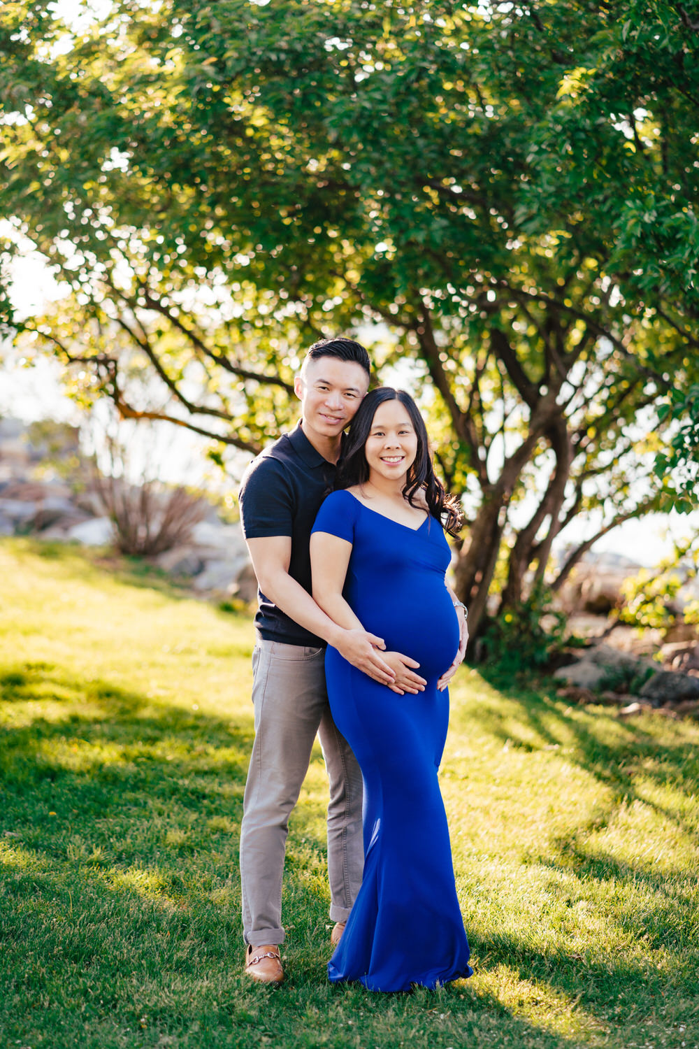a pregnant couple standing in the grass in front of a tree.