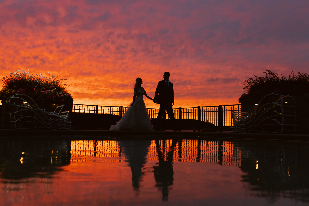 A bride and groom standing next to a pool at sunset.